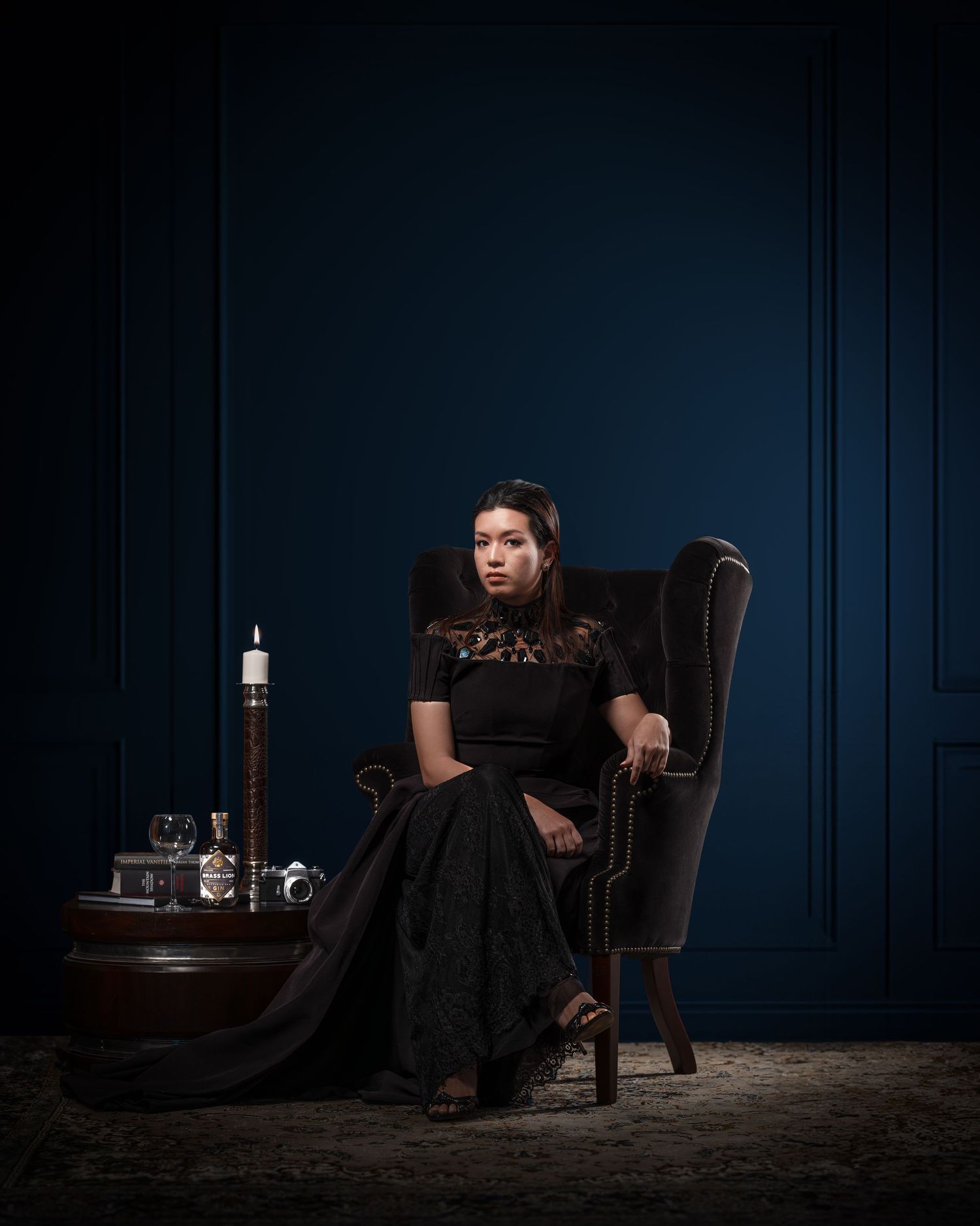 Woman in elegant black dress seated in velvet armchair against deep blue wall with candle and decor.
