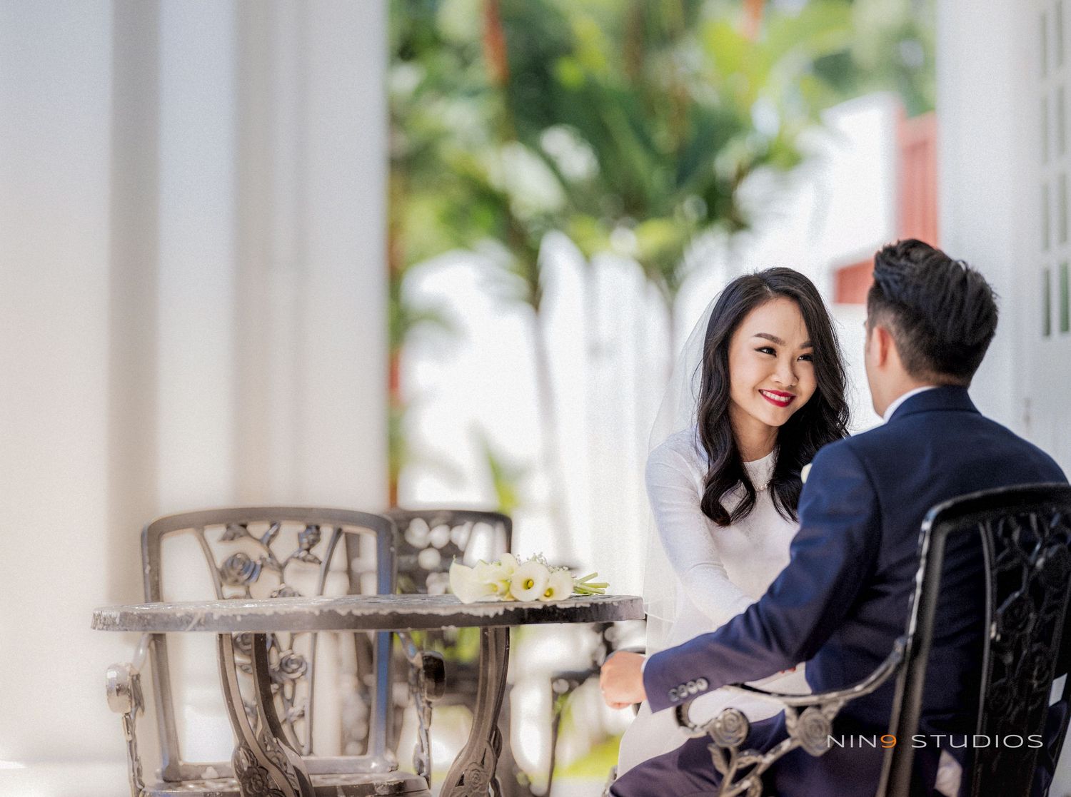 Couple sharing a romantic moment at an outdoor table, woman in white dress smiling at man in navy suit.