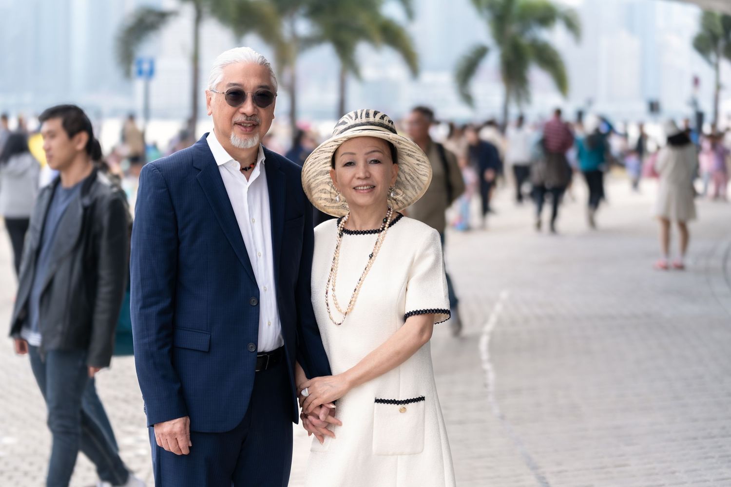 Elderly Asian couple holding hands and smiling on a busy waterfront promenade, dressed in elegant white and navy attire.