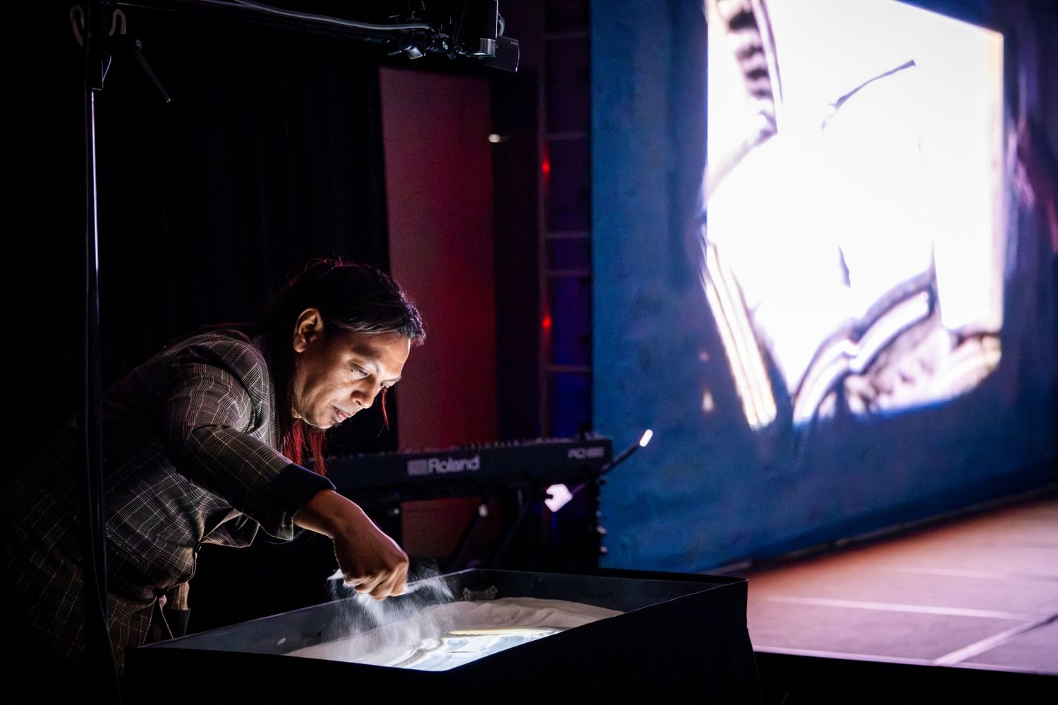 Musician performing on a Roland keyboard with a large projection screen displaying visuals in a darkened venue.