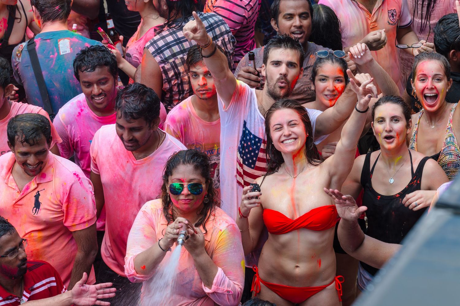 Crowd celebrating Holi festival with colorful powder, woman holding American flag.
