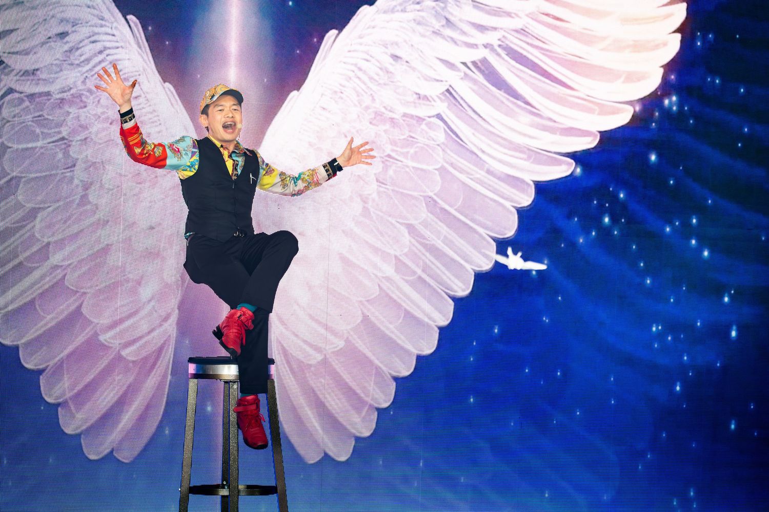 Entertainer posing on stool in front of large angel wings stage backdrop.