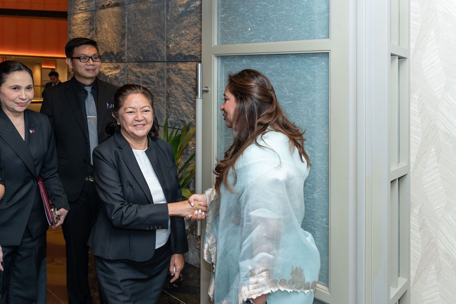 Two women in formal attire shake hands at a doorway, greeted by colleagues in a professional setting.