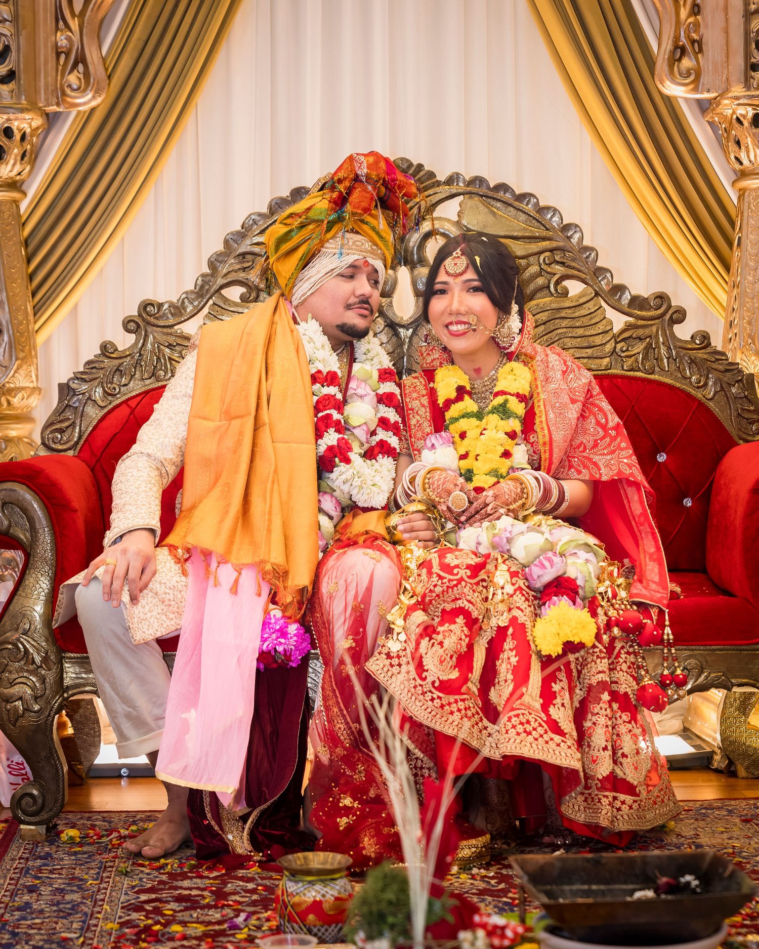 Couple in traditional Indian wedding attire seated on red sofa with floral garlands and decorations.