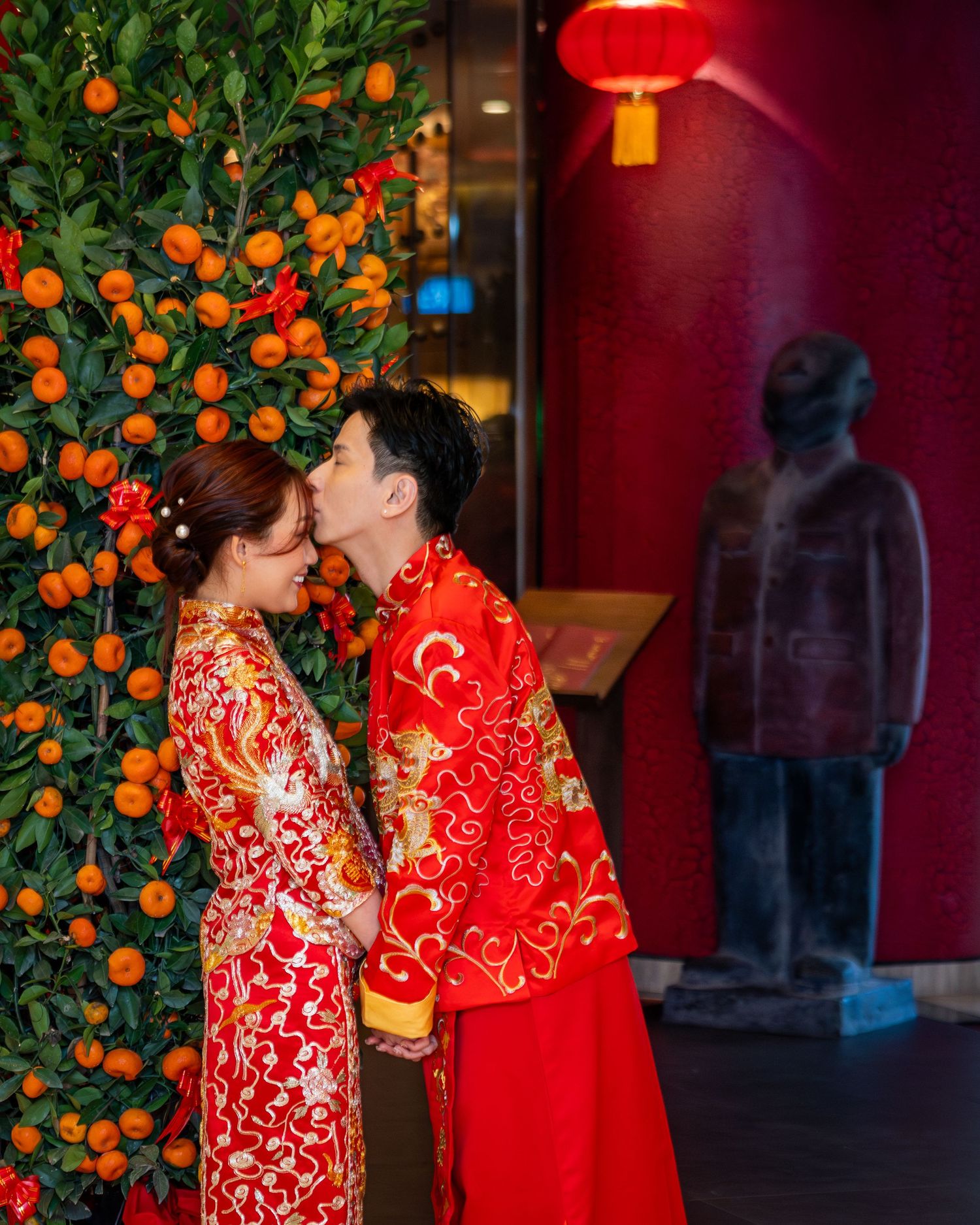 Chinese couple in traditional red wedding attire kissing beside an orange tree with red lantern decor.