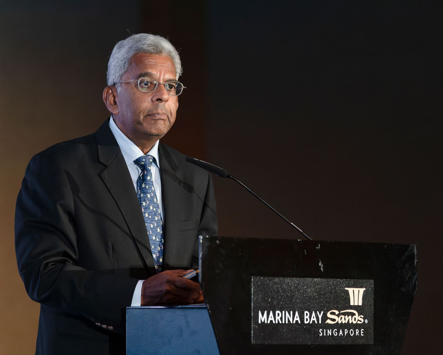 Senior businessman in suit speaks at Marina Bay Sands podium during formal Singapore event.