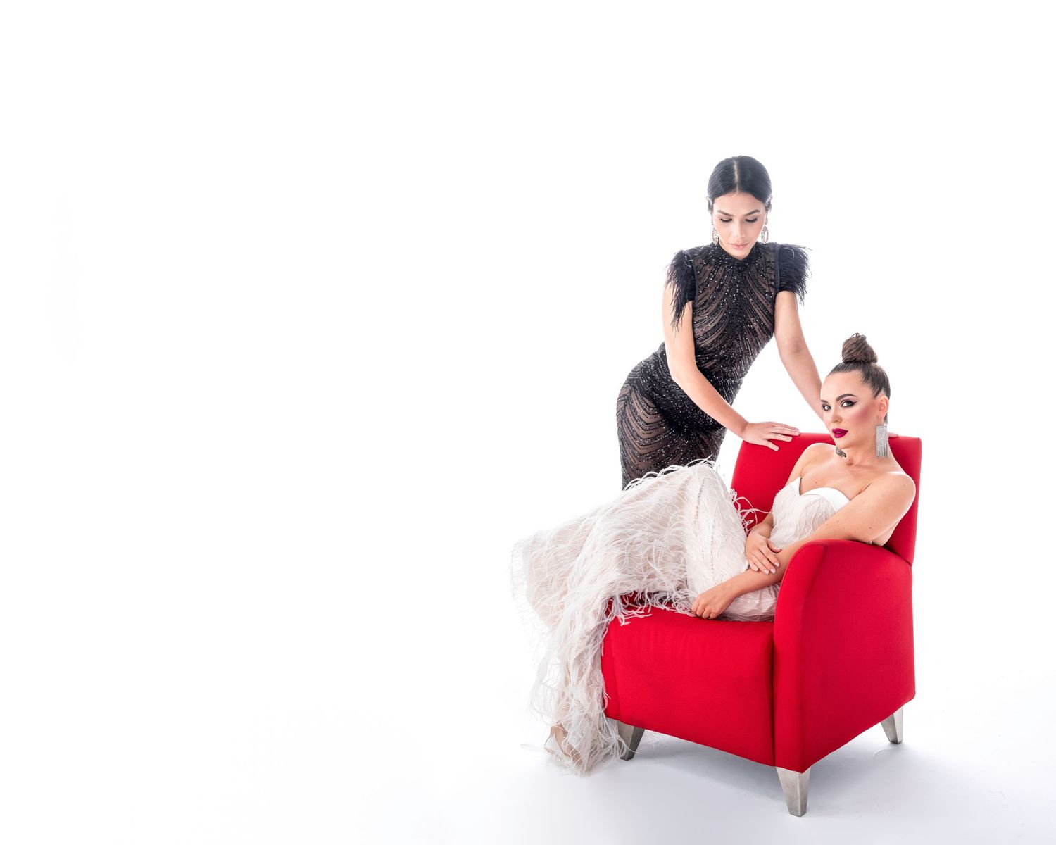 Two elegant women pose with a red armchair, one in black dress standing, one in white gown reclining, on white background.