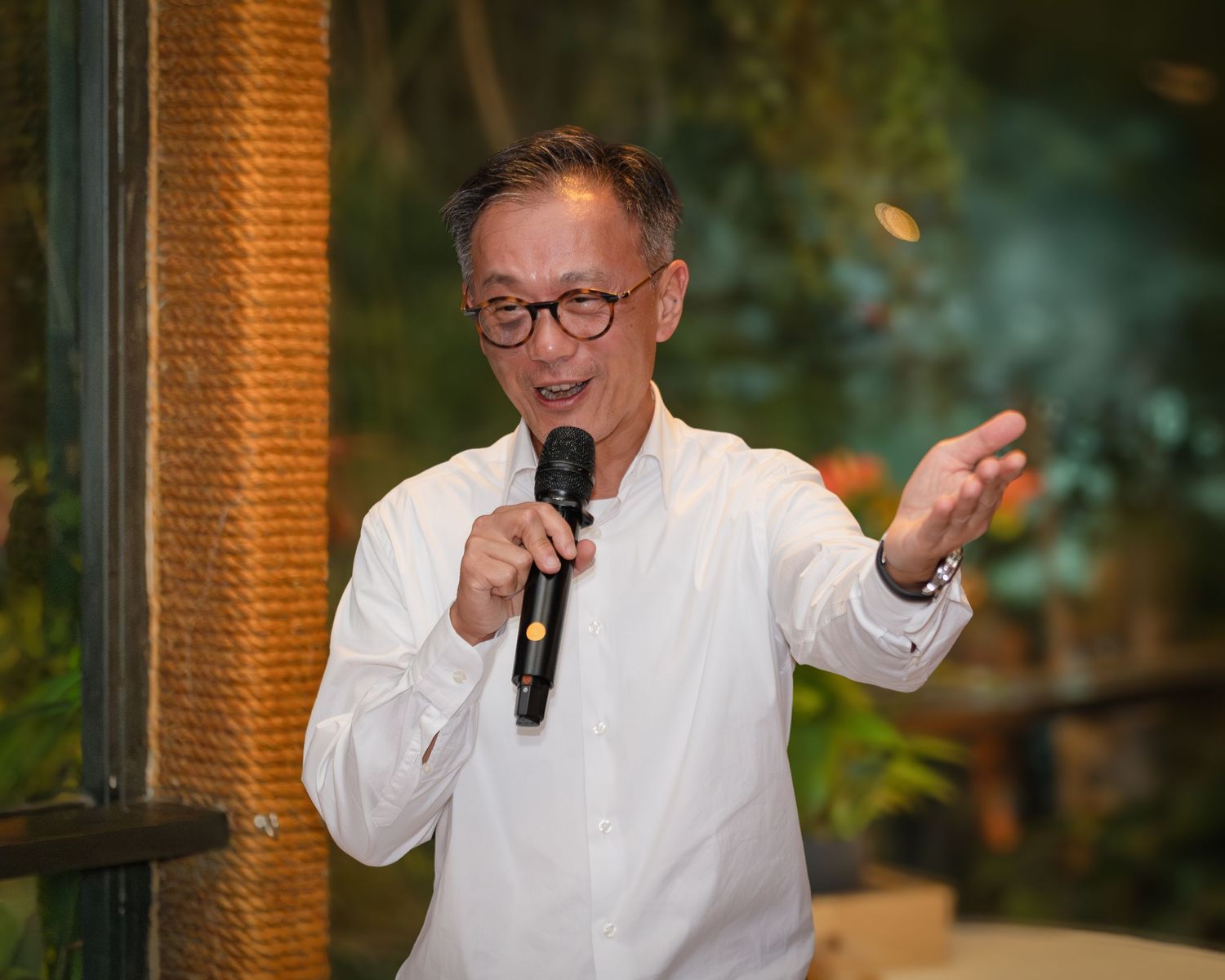 Middle-aged Asian man in white shirt speaking into microphone at indoor event, gesturing with hand.