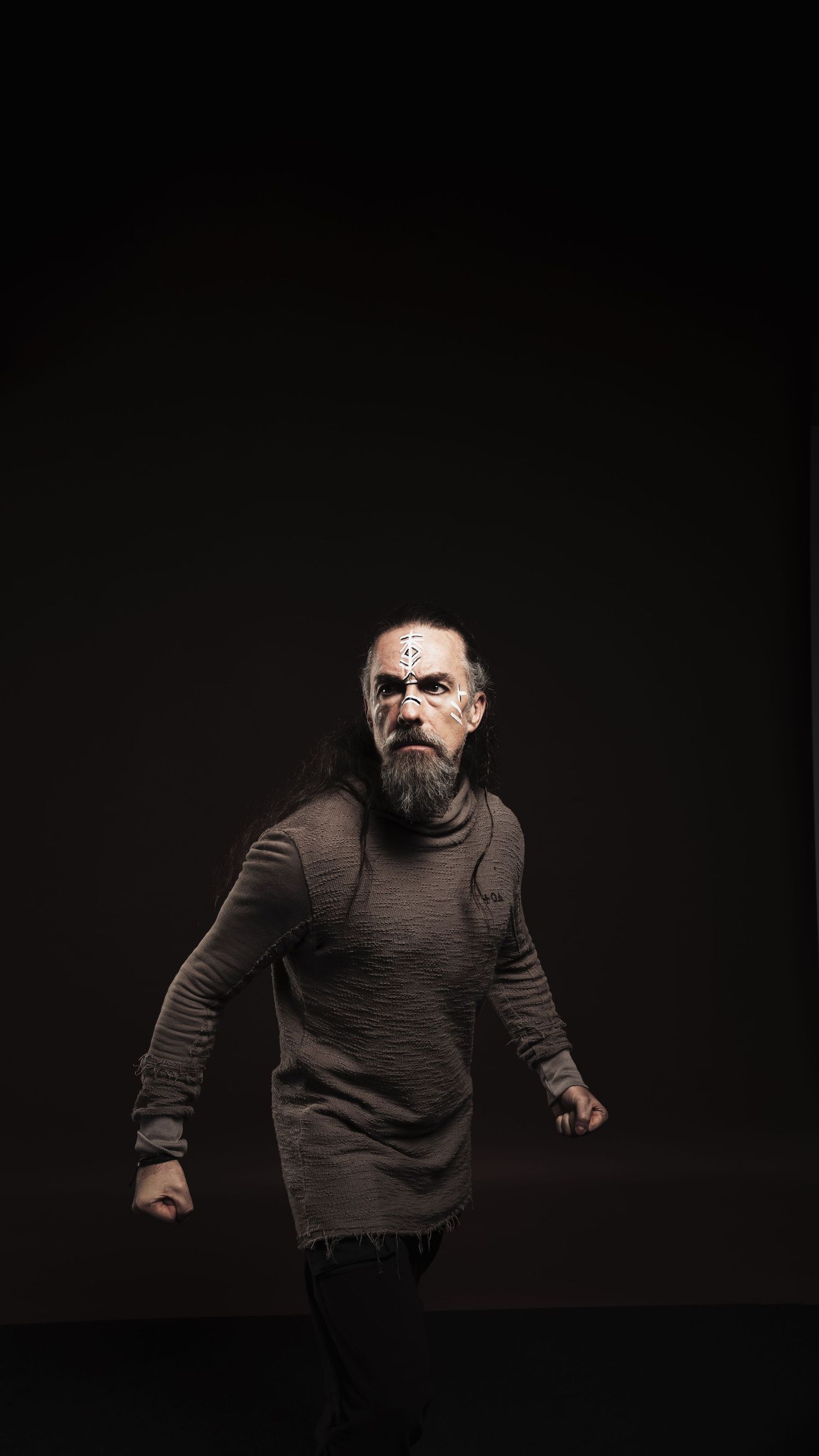 Bearded man with face paint in dark sweater poses dramatically against black background.