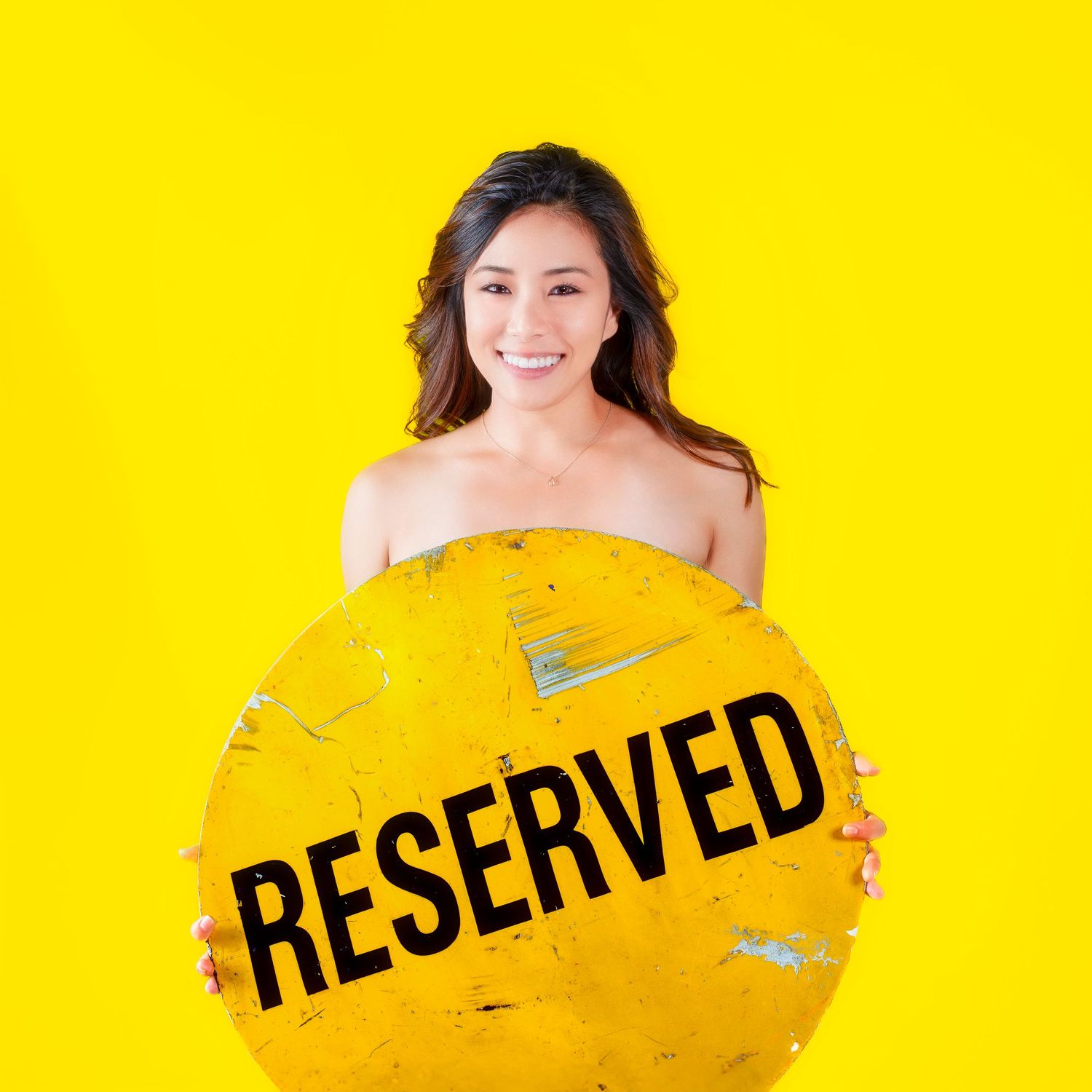 Smiling woman holding a round yellow reserved sign against a bright yellow background.