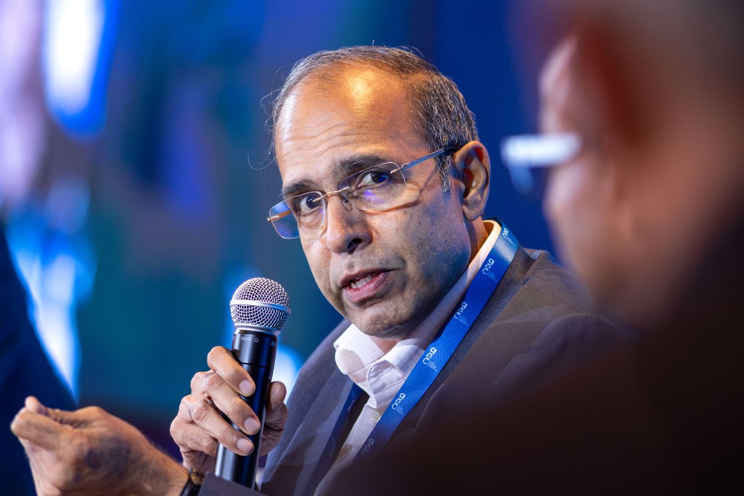 A man in a dark blazer speaks into a microphone during a professional conference panel discussion.