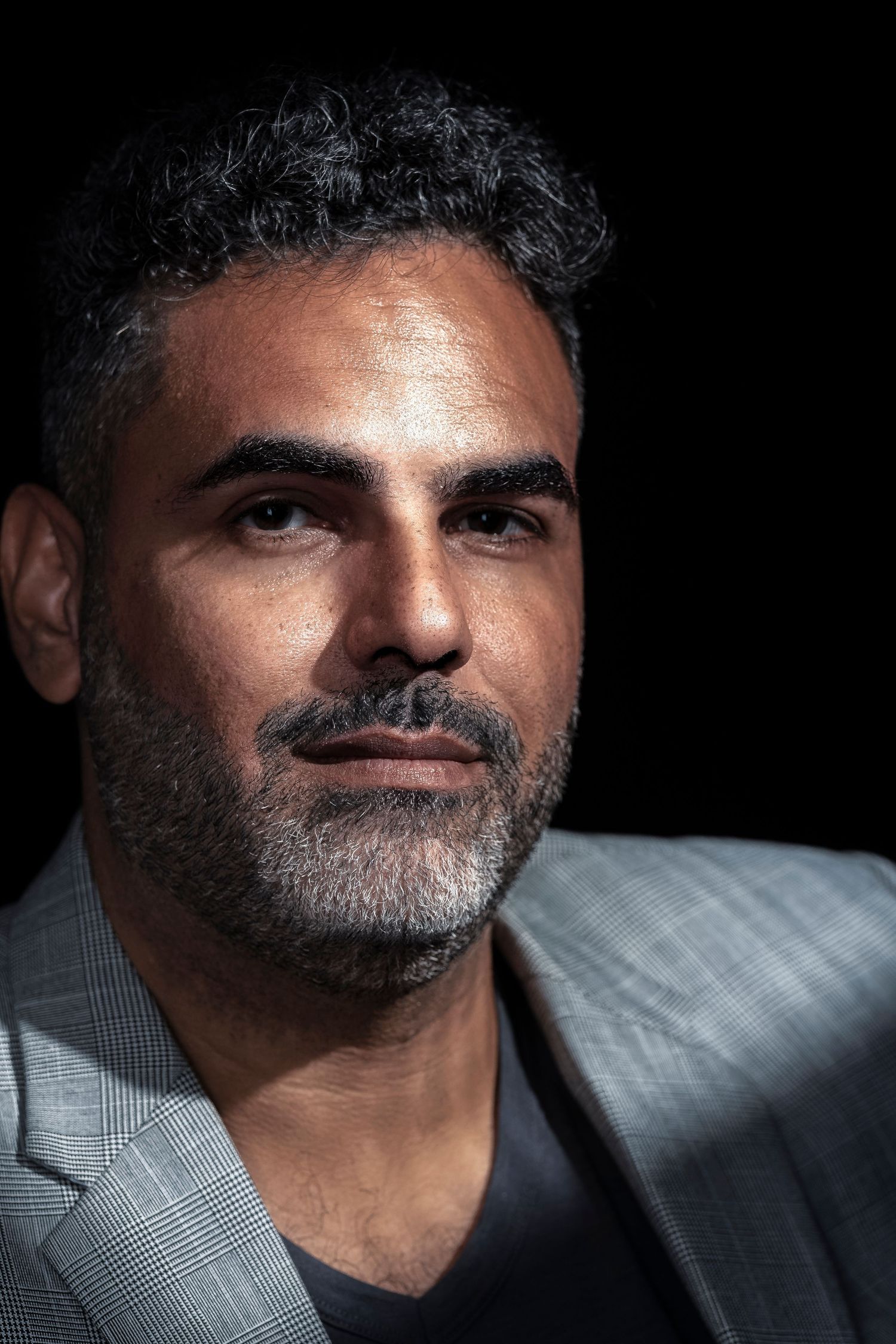 A middle-aged man with dark curly hair and a short beard wearing a gray blazer against a dark background.