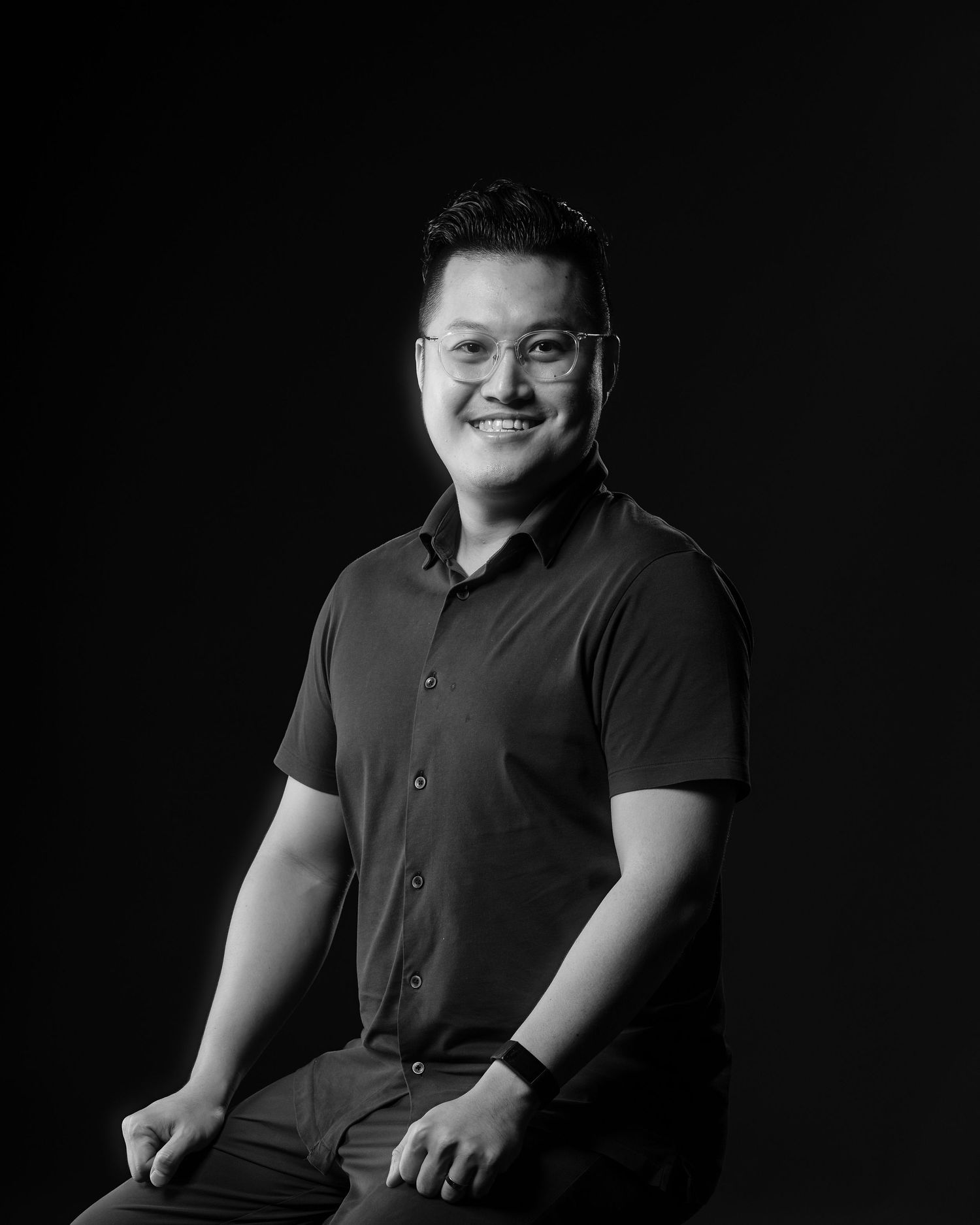 Smiling Asian man in dark shirt poses for black and white professional portrait photo.