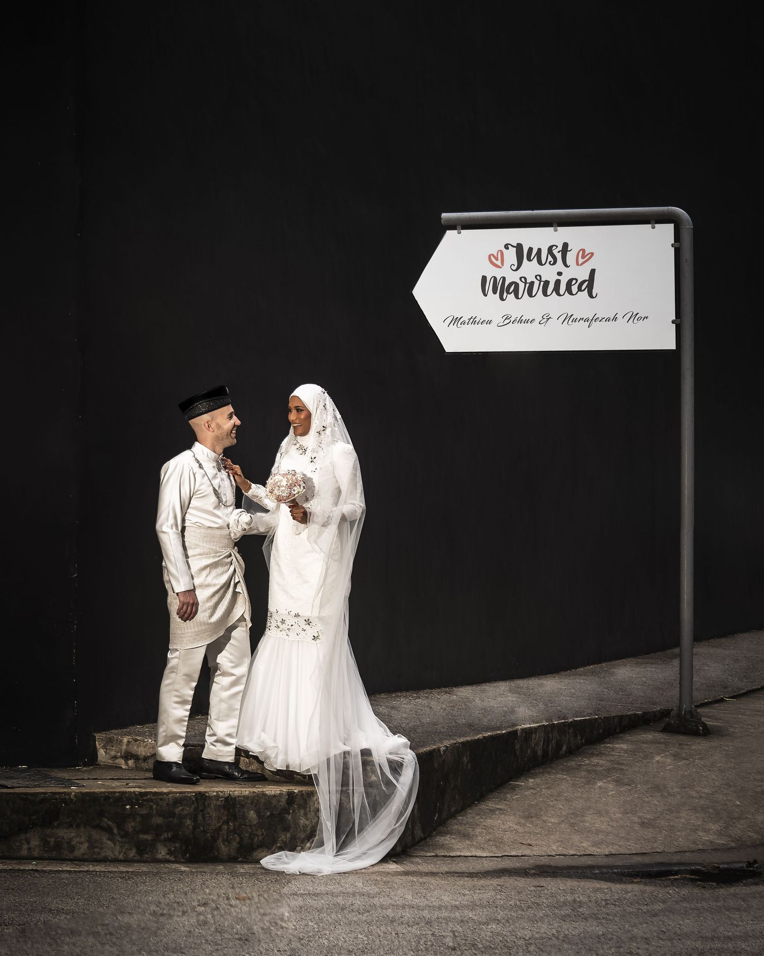 Newlywed couple in white wedding attire posing beside a Just Married sign outdoors at night.