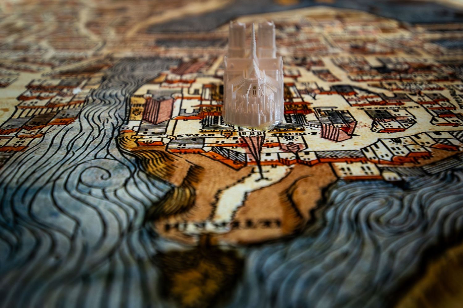 A detailed illustrated antique city map with a small clear figurine placed on top, shown in shallow depth of field.