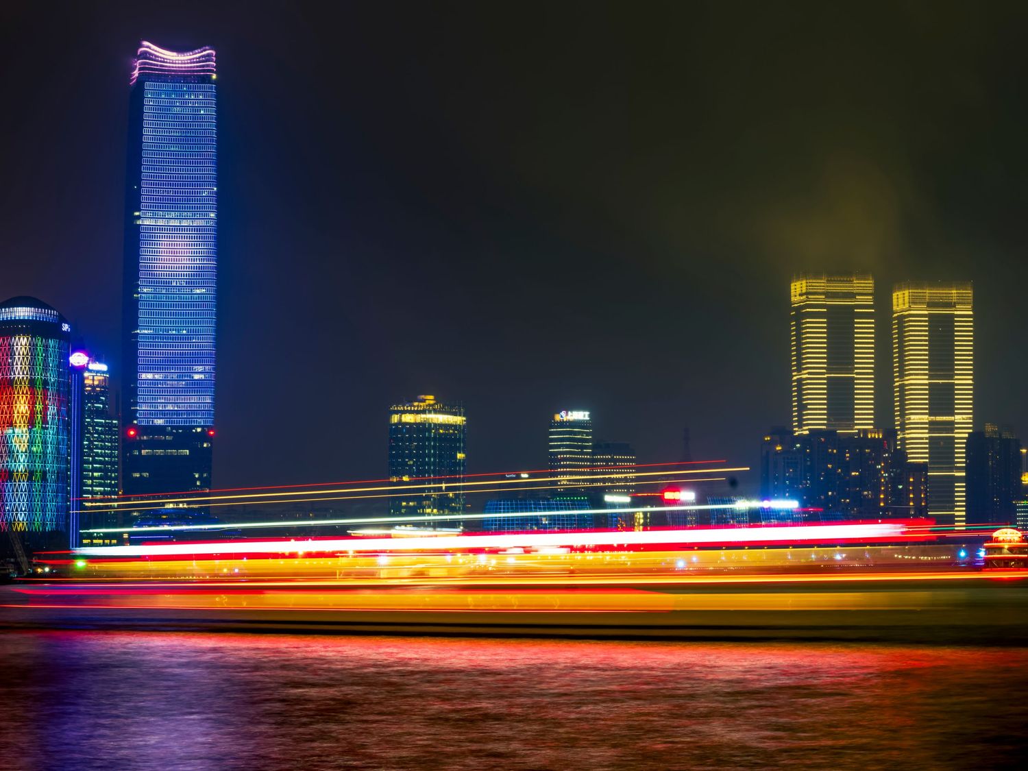 Night cityscape with colorful light trails over water, illuminated skyscrapers glowing against dark sky.