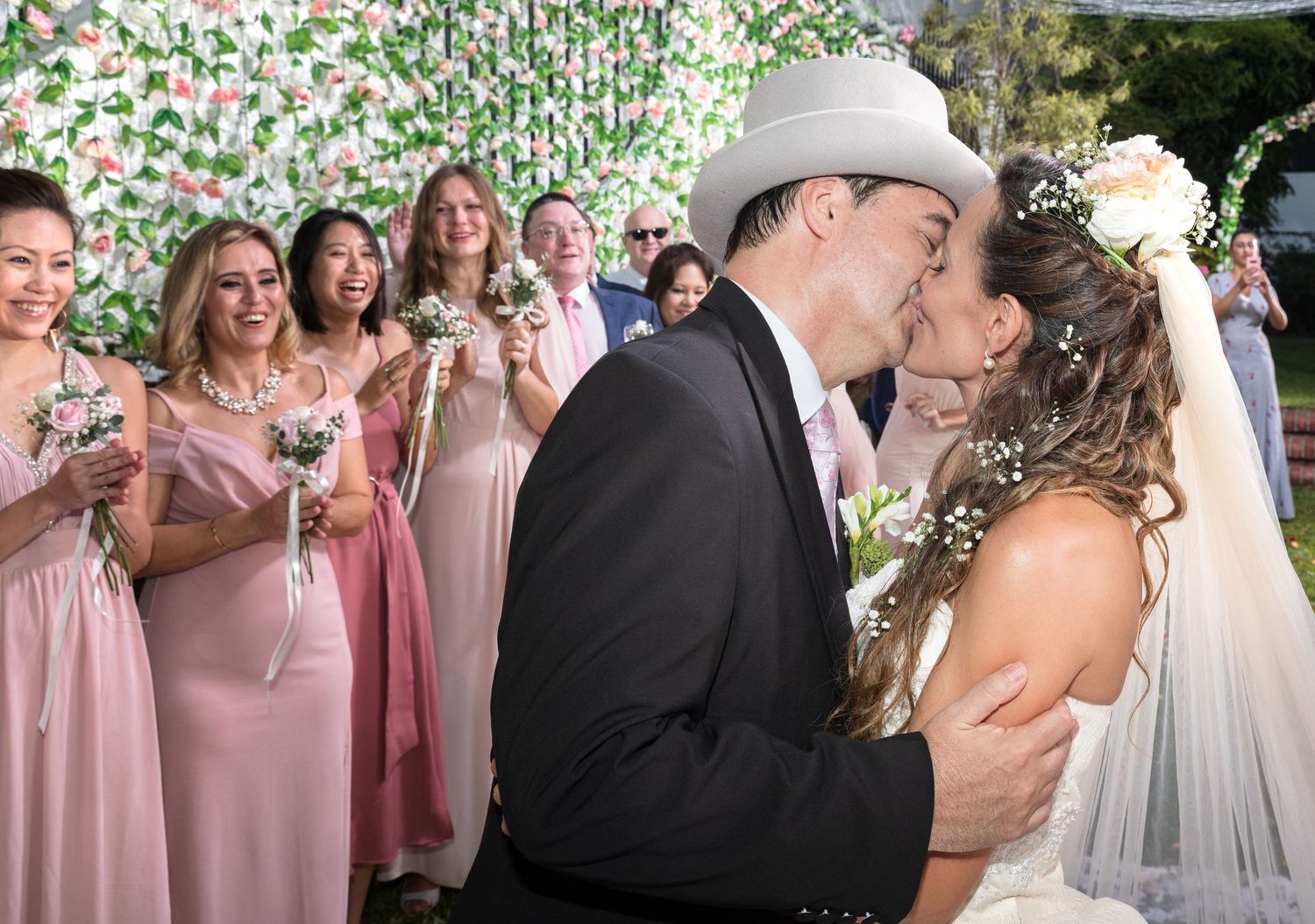 Bride and groom share first kiss at outdoor wedding ceremony with bridesmaids in pink dresses and floral backdrop.