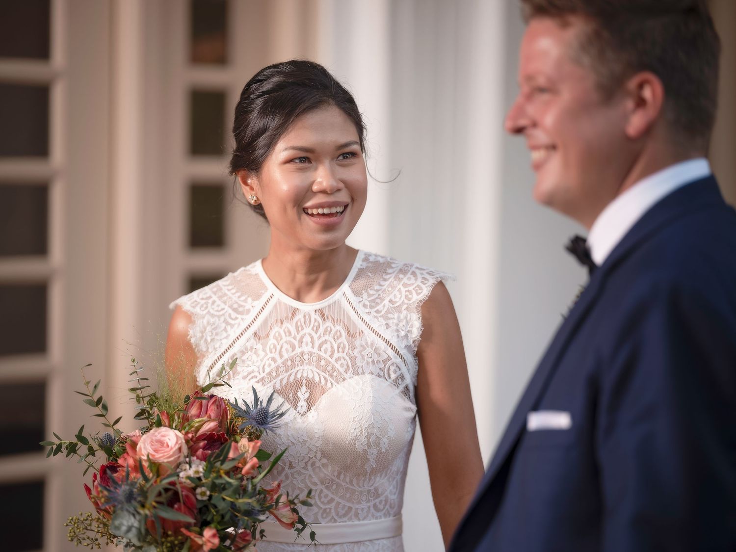 Smiling bride in lace wedding dress holding floral bouquet facing groom in navy suit outdoors.
