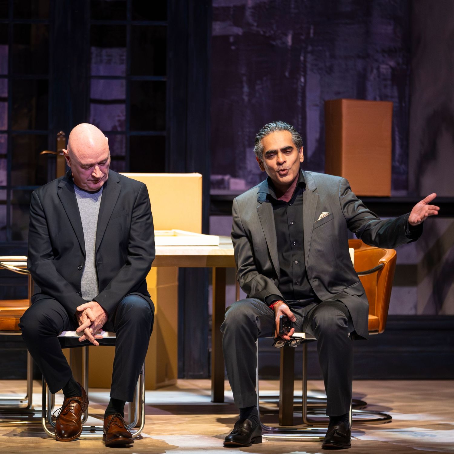 Two men seated on stage during a formal panel discussion or theatrical performance in a dimly lit venue.