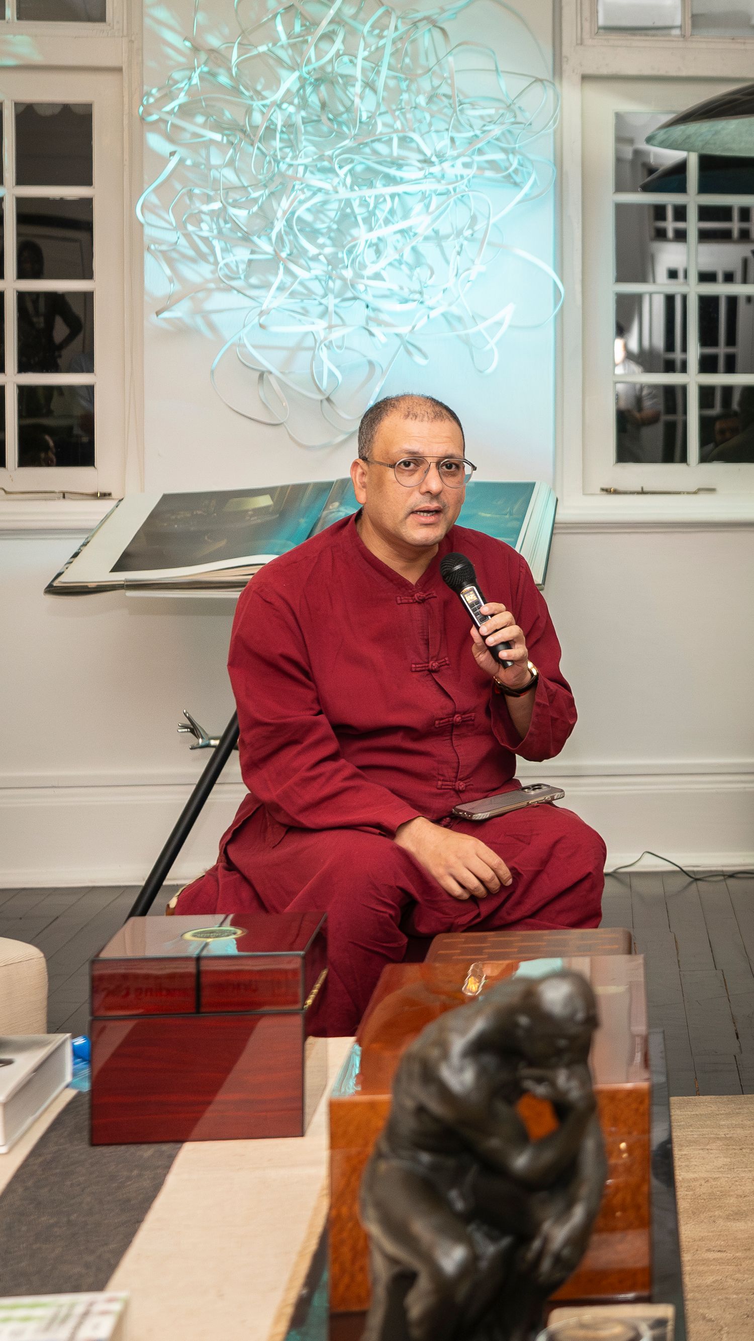 Man in red robes speaks into microphone while seated, with glowing art installation backdrop.