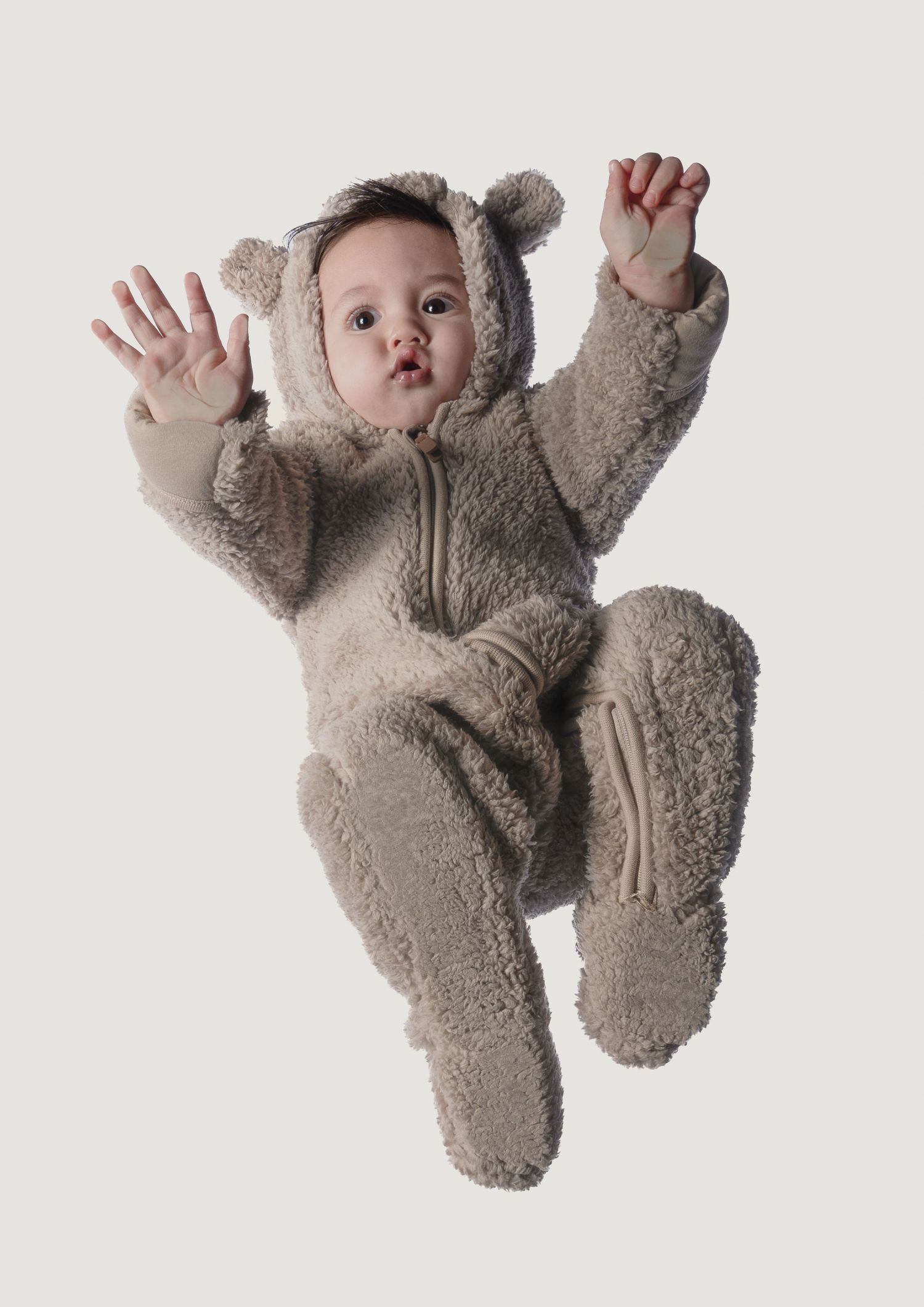 Baby in a cozy beige teddy bear fleece onesie with ears, arms raised, isolated on light background.