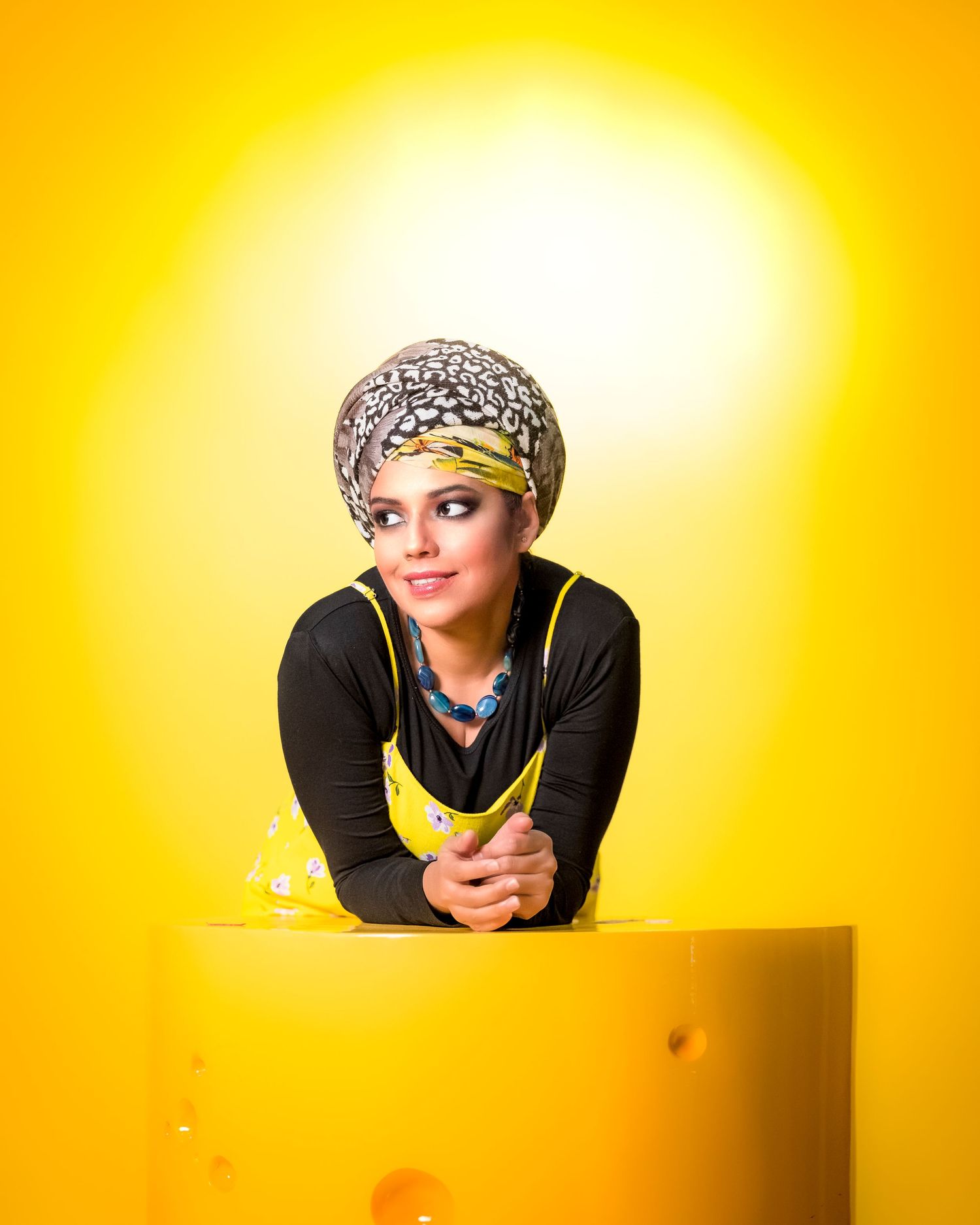 Young woman in colorful head wrap and yellow apron posing against vibrant yellow background.