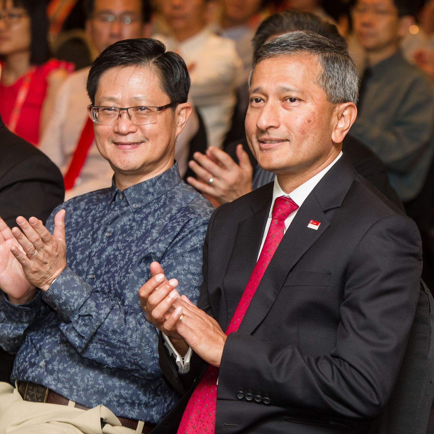 Two formally dressed men applaud at an official ceremony, one wearing a Singapore flag pin on his lapel.