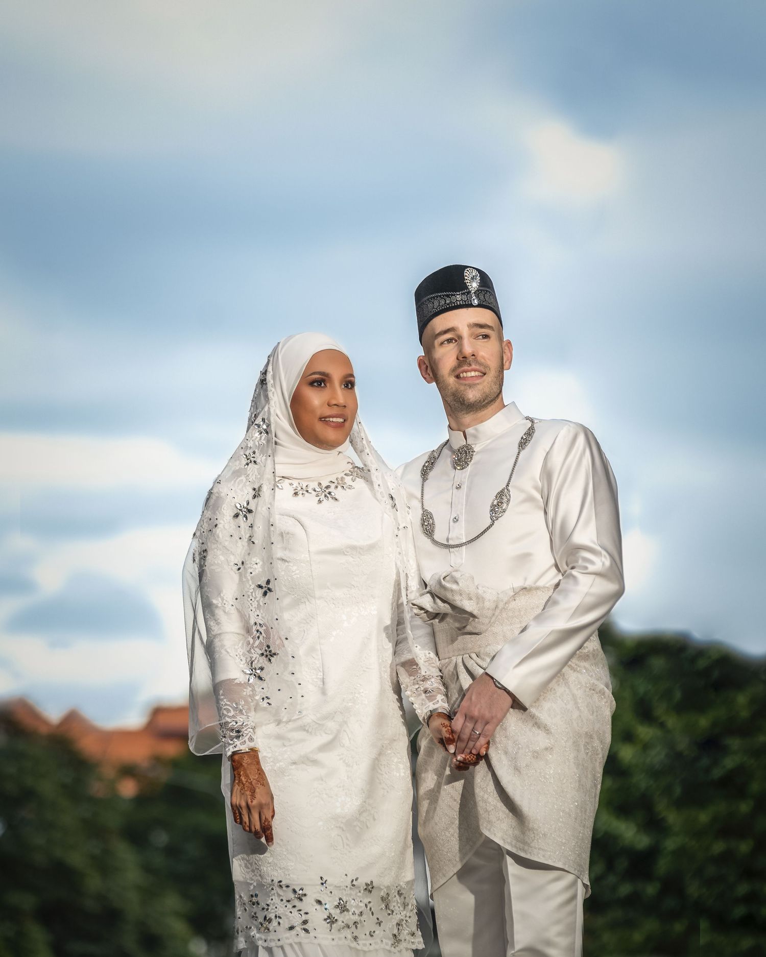 Muslim wedding couple in white traditional Malay attire posing outdoors under a blue sky with lush greenery.
