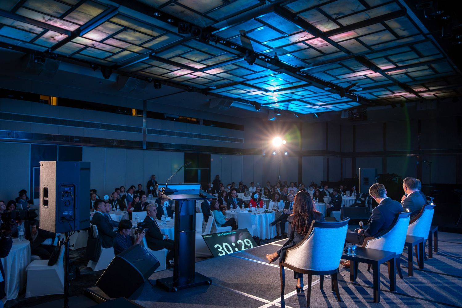 Large conference audience seated in a dimly lit ballroom during a panel discussion on stage.