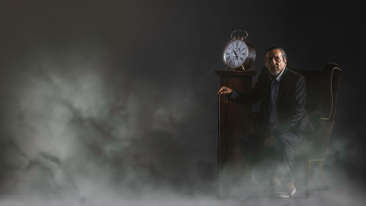A mysterious man in a suit stands beside a grandfather clock surrounded by dramatic fog in a dark studio setting.