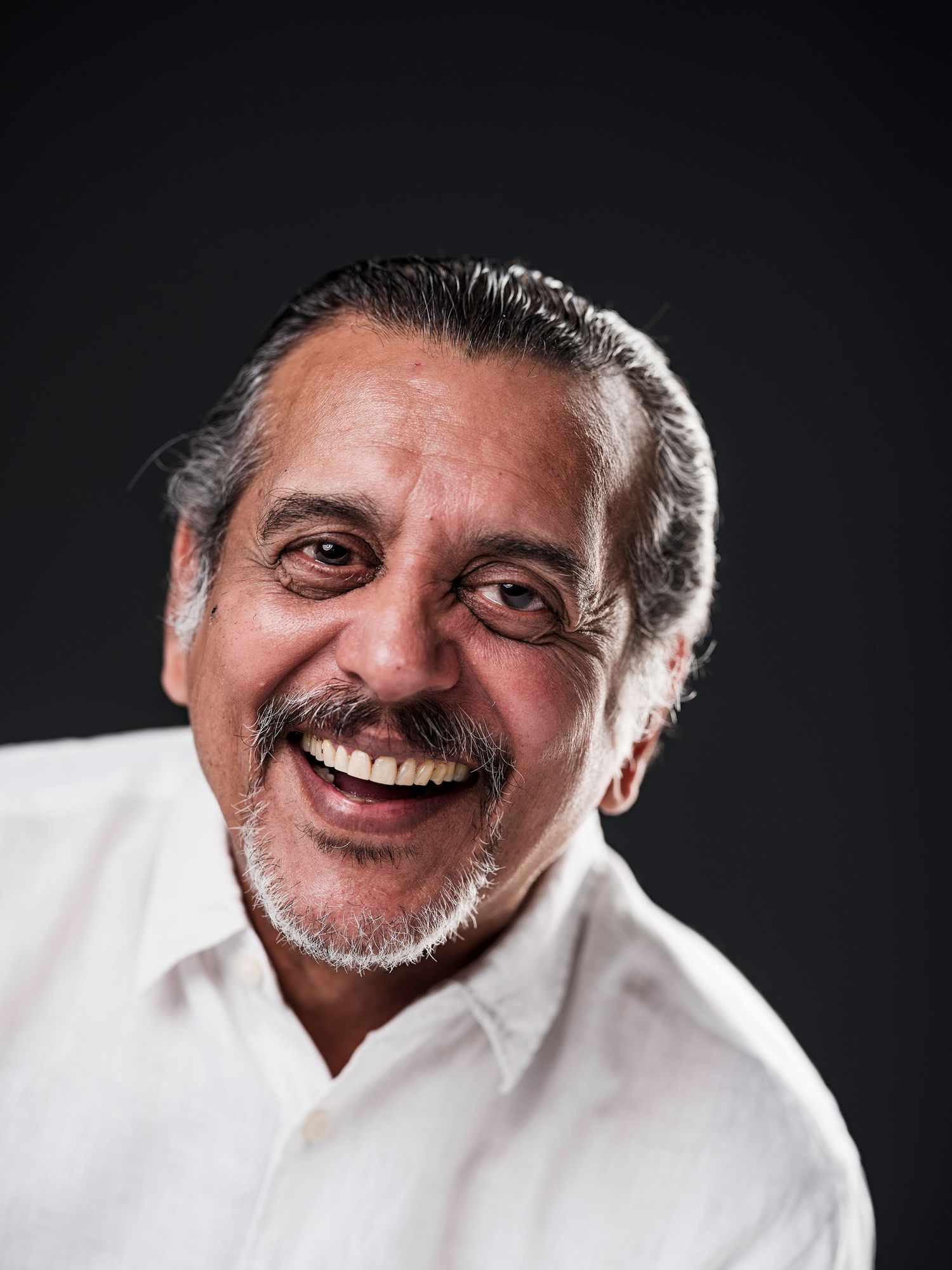 Smiling middle-aged man with gray hair and beard wearing white shirt against dark background.