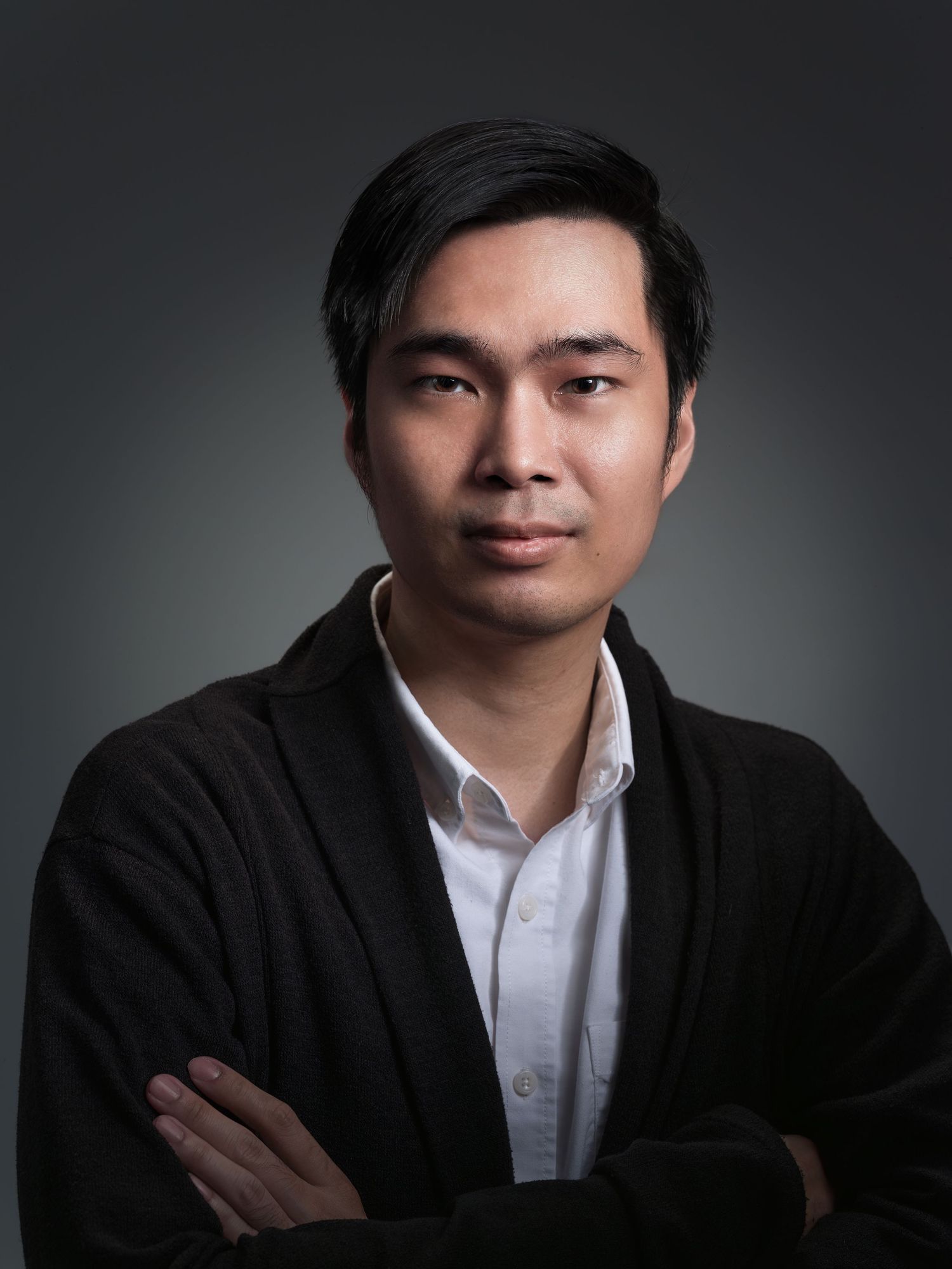 A young Asian man with crossed arms wearing a black cardigan over a white shirt poses for a professional headshot.