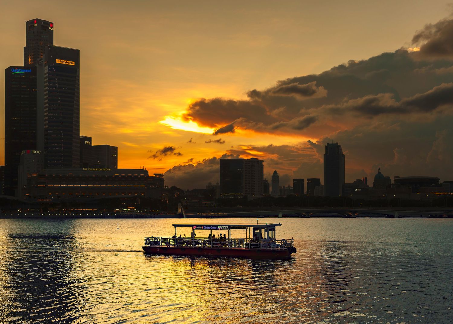 A boat silhouetted against a golden sunset over a city harbor with dramatic clouds and glowing water reflections.