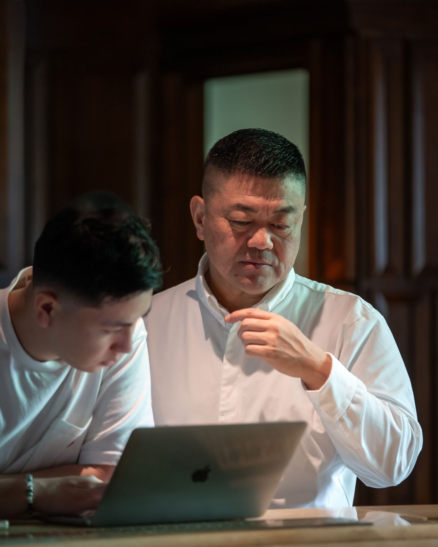 Two men in white shirts collaborate over an Apple laptop in a dimly lit wooden room.
