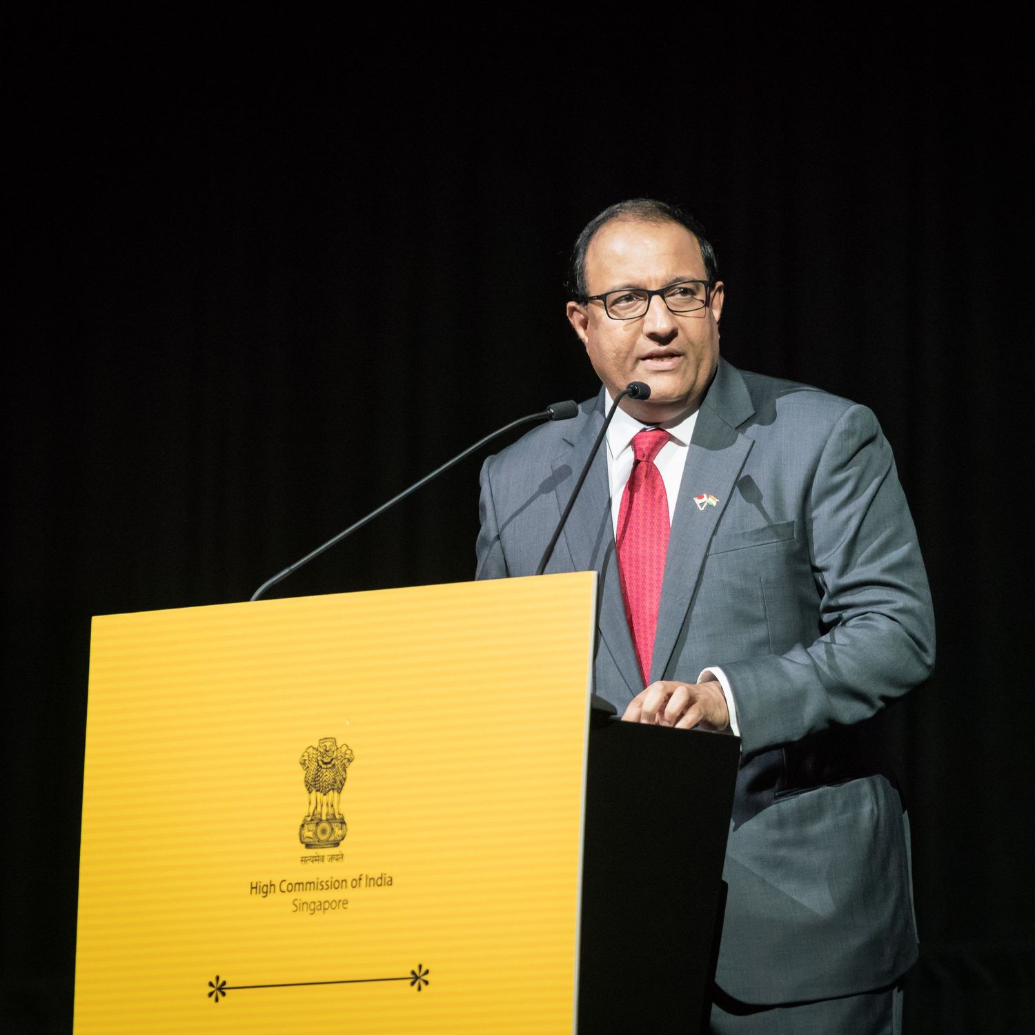 A man in a grey suit with red tie delivers a speech at a podium bearing the High Commission of India Singapore seal.