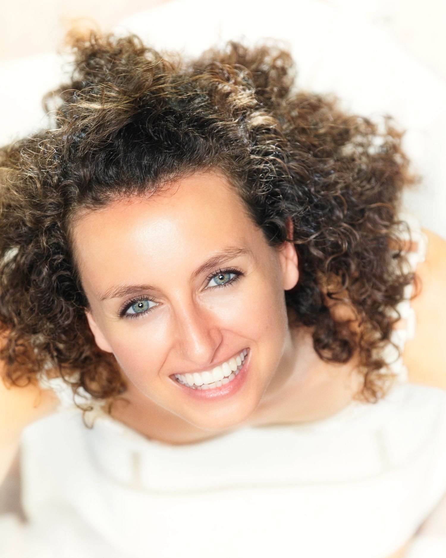 Smiling woman with curly brown hair and blue eyes, wearing white, photographed from above in bright lighting.