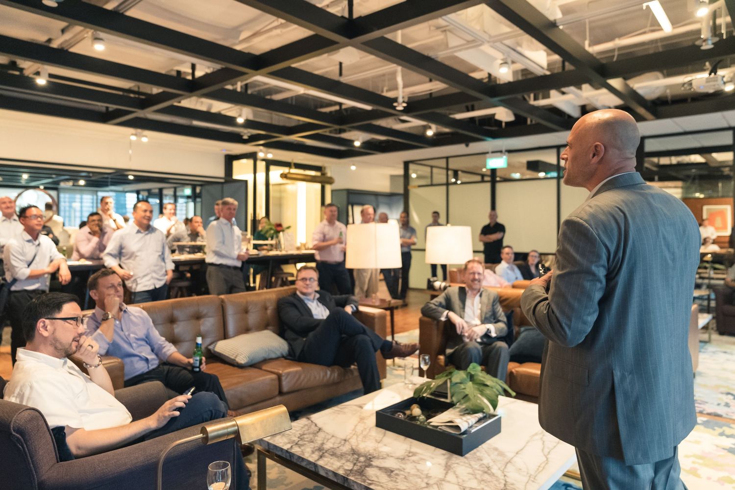 Business professional addresses a group of colleagues seated on sofas in a modern coworking space.