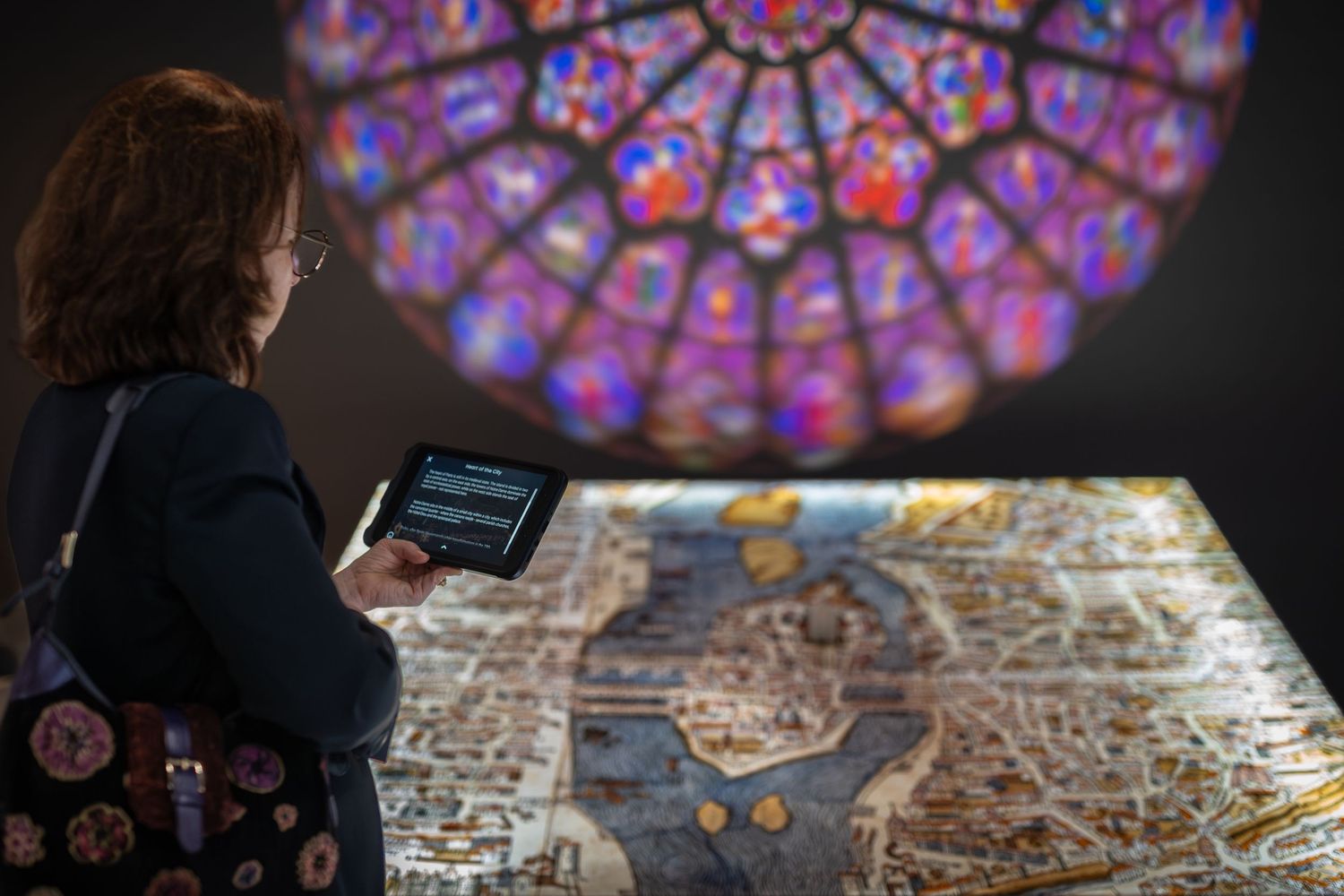 Woman uses tablet to explore illuminated medieval map display beneath colorful rose window projection in museum.