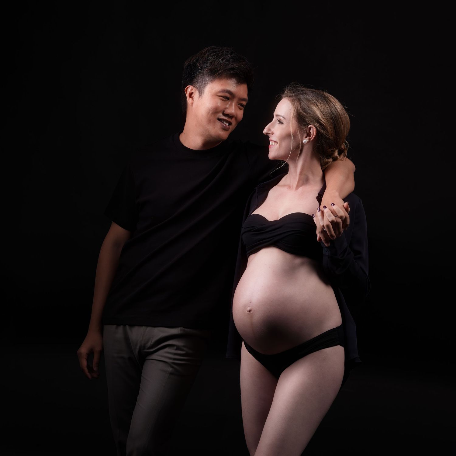 A couple poses for a maternity photo shoot against a dark background, showcasing the woman's baby bump.