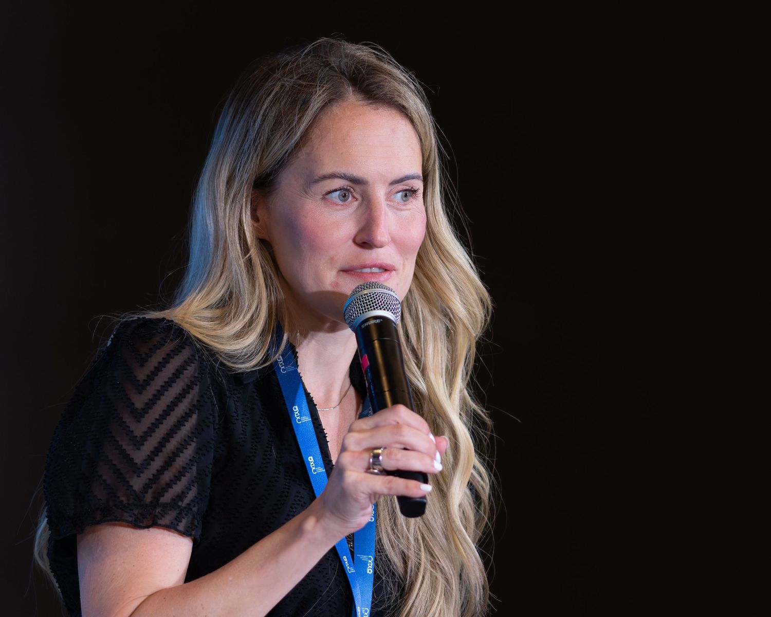 A blonde woman in a black top speaks into a microphone wearing a blue lanyard at a conference.