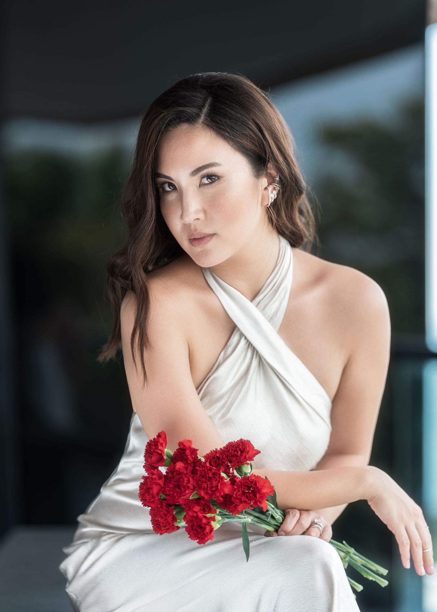 Woman in white halter dress holding red carnations, seated elegantly with soft bokeh background.