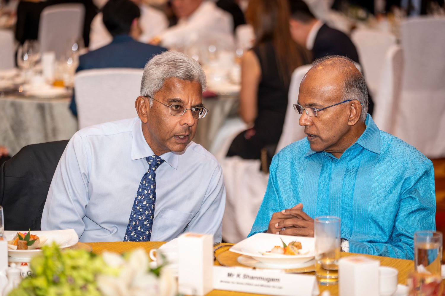 Two men in formal attire engage in conversation at a banquet dinner table with place settings and name cards.