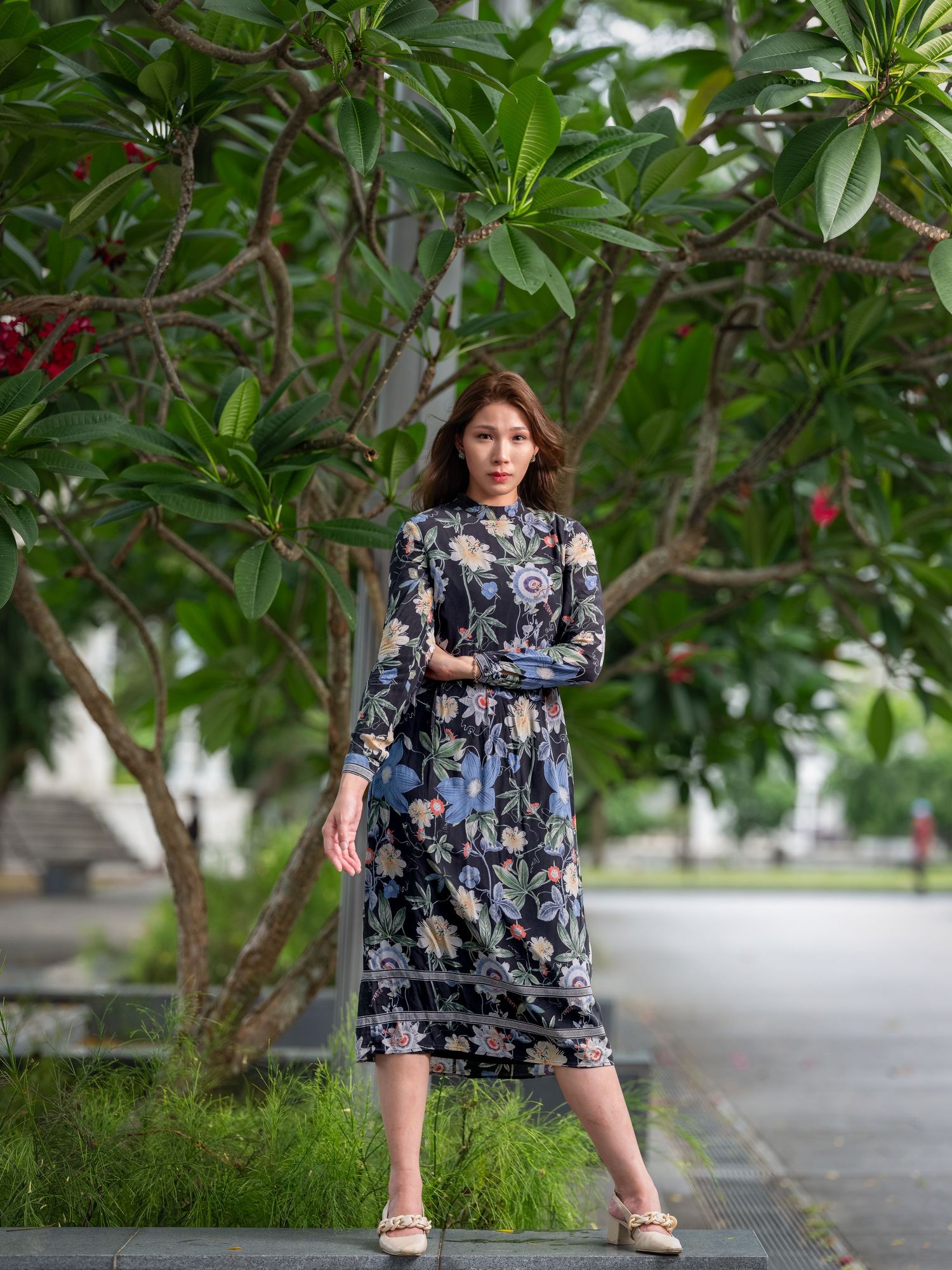 A woman in a floral midi dress poses outdoors near lush green trees on a sunny day.