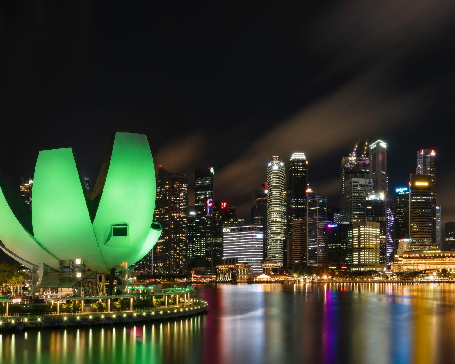 Singapore skyline at night with illuminated green ArtScience Museum lotus flower and colorful city reflections on Marina Bay.