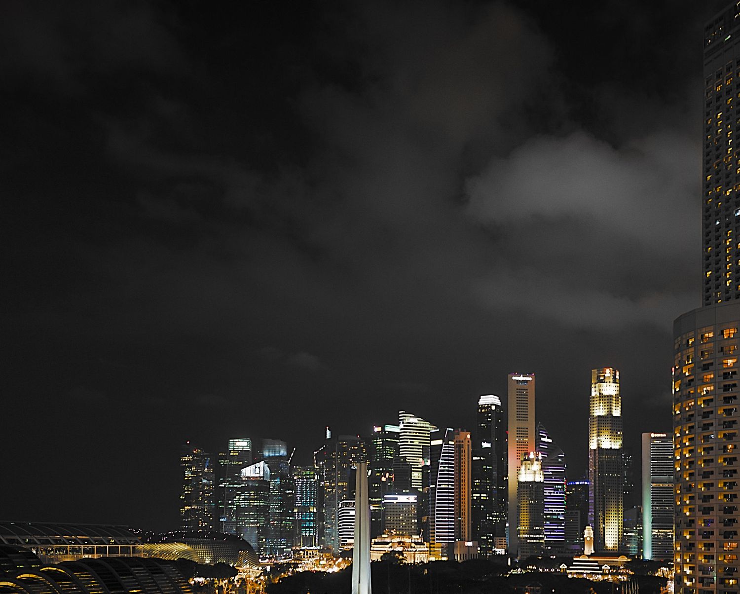 A dramatic nighttime skyline of Singapore with glowing skyscrapers against dark storm clouds over the city.