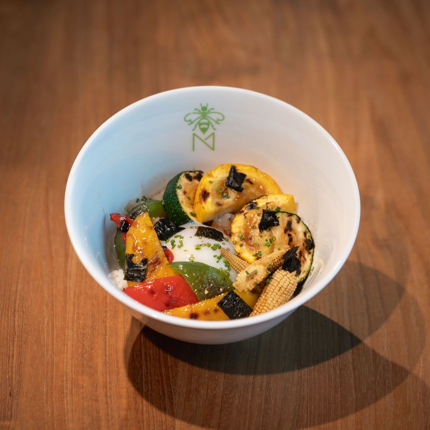 Grilled mixed vegetables in a white bowl with a bee logo on a wooden table.