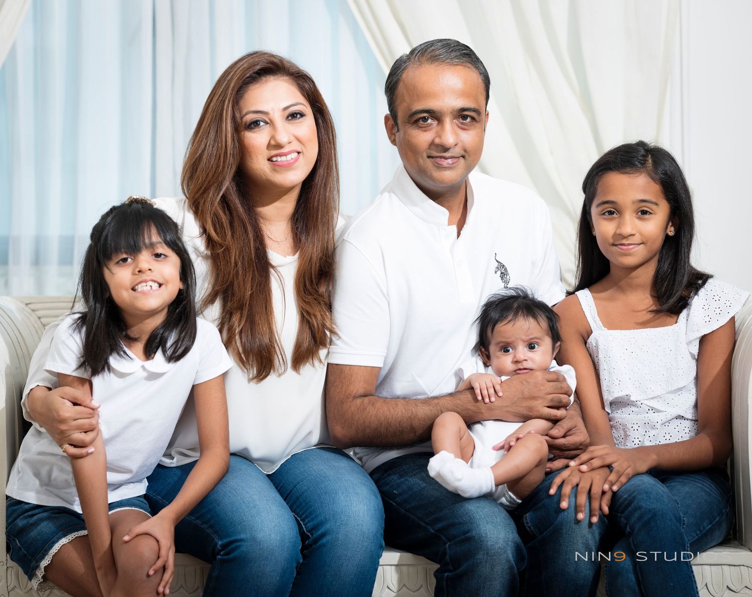 Family portrait of parents with three children, dressed in white tops and jeans, posing together on a sofa.