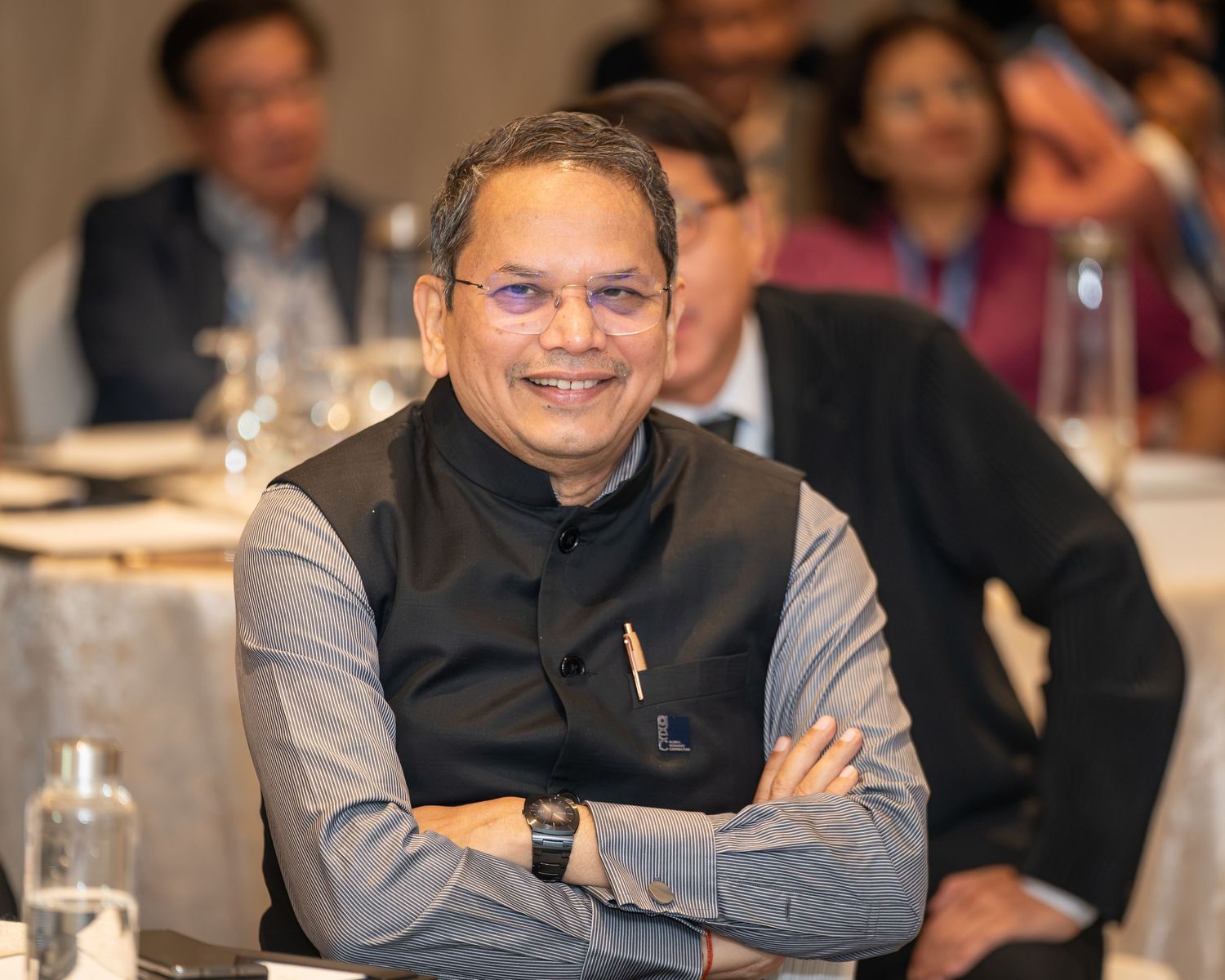 Smiling South Asian man in black vest sits with arms crossed at a formal conference event.