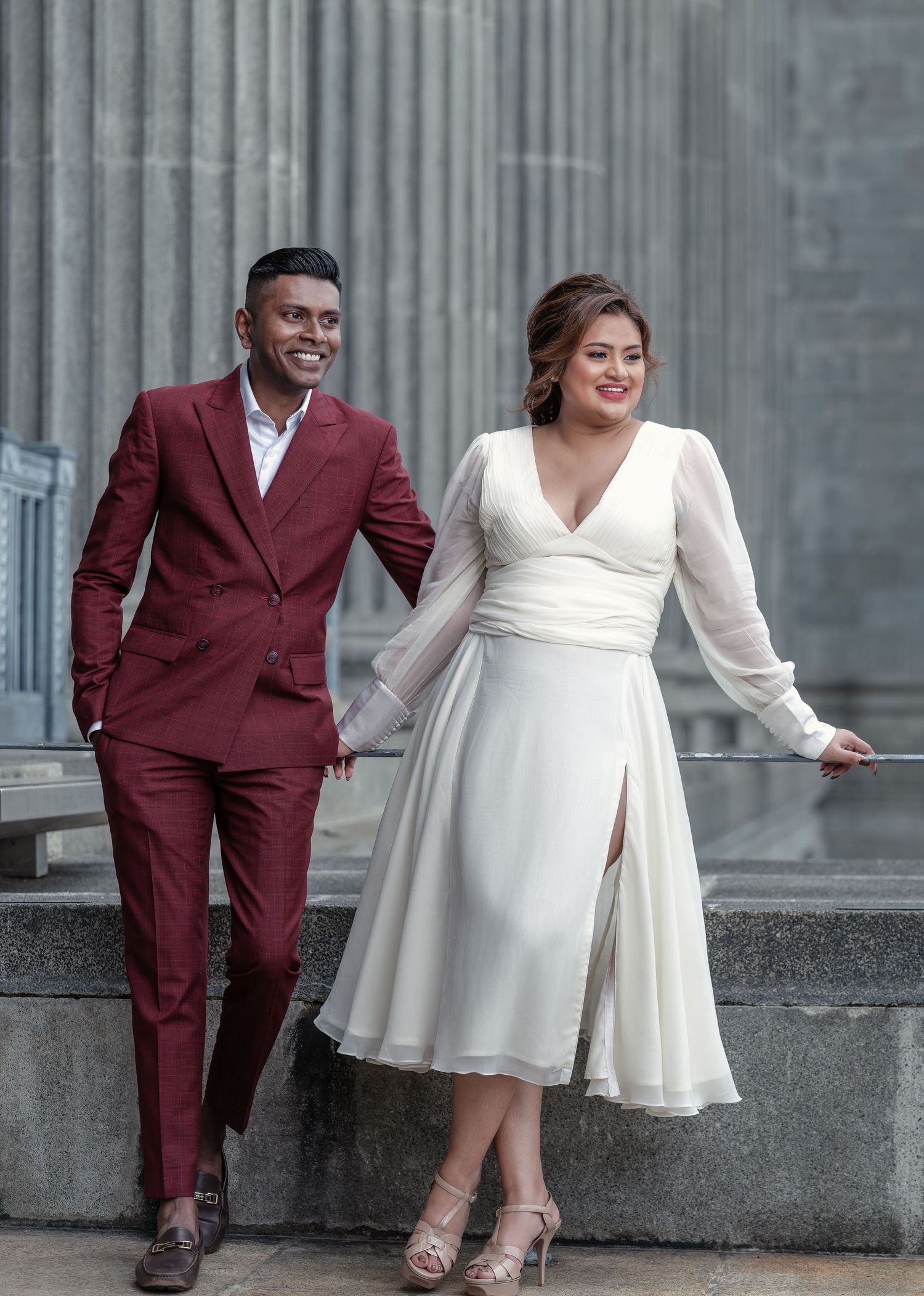 Stylish couple walking together, man in burgundy suit and woman in white midi dress, posing near stone columns.
