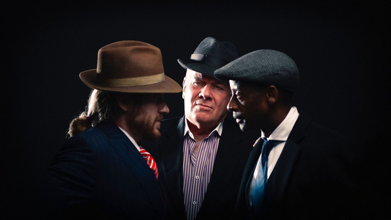 Three men in stylish hats and suits pose dramatically against a dark background in a moody, cinematic portrait.